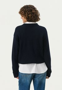 Navy knitted sweater with a cropped design, ribbed edges, and cable knit detailing on sleeves, worn over a white shirt and denim jeans.