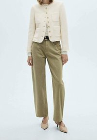 Cremfarbener Texturjacke mit goldenen Knöpfen und zwei Taschen, kombiniert mit hoch taillierten olivgrünen Hosen und beigen spitzen Schuhen.