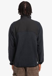 Black fleece jacket with a textured finish, featuring a smooth black panel across the upper back, and ribbed cuffs.