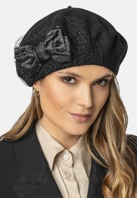 Béret en laine noir avec un grand nœud en satin et des détails en filet. Présente de petites perles décoratives et une texture douce. Forme ronde classique.