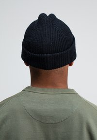 Gehaakte beanie in donkermarine met een geribbelde textuur en twee opvallende naden op de kroon; gedragen met een groene sweatshirt.