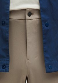 Beige, srednje teški materijalni hlače s ravnim prednjim dijelom, sa malim crnim dugmetom i bočnim šavom. Upotpunjene plavim odijelom s dugmadima.