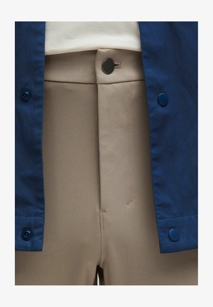 Beige, srednje teški materijalni hlače s ravnim prednjim dijelom, sa malim crnim dugmetom i bočnim šavom. Upotpunjene plavim odijelom s dugmadima.