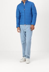 Blauwe pufferjack met horizontale quilt, hoge kraag en een ritssluiting aan de voorkant. Gepaard met lichtblauwe jeans en witte sneakers.