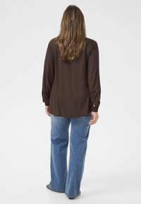 Brun långärmad skjorta med lös passform, mjukt material och knäppning vid ärmen, matchad med ljusblå utsvängda jeans och bruna skor.