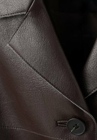 Brązowa skórzana marynarka o teksturowanym wykończeniu, z wyraźnie zaznaczonymi klapami i pojedynczym zapięciem na guzik. Szwy zdobiące krawędzie.