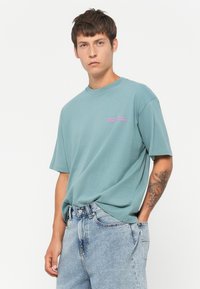 Light teal cotton t-shirt with short sleeves and a crew neckline, featuring pink text on the left chest. Worn with light blue denim shorts.