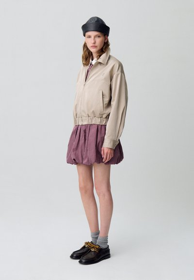 Giacca bomber beige con vita elastica, abbinata a una gonna a palloncino in gingham viola. Il modello indossa calzini grigi e scarpe scure con dettagli dorati.