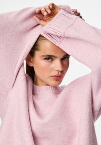 Rózsaszín kötött pulóver, lekerekített nyakkivágással, bő ujjakkal és texturált részletekkel a mandzsettánál. Puha, bolyhos anyag ad mélységet a megjelenésnek.