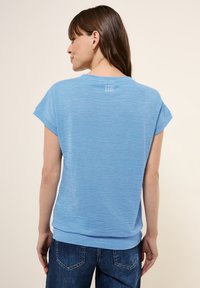 T-shirt bleu clair à manches courtes avec une finition texturée. Il présente un col rond et un petit logo blanc au dos près du col.