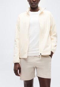 Άτομο που φορά ανοιχτό μπεζ ζακέτα με φερμουάρ, λευκό πουκάμισο και μπεζ σορτς, στέκεται μπροστά σε απλό λευκό φόντο.