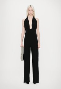 Salopetă neagră cu gât halter și decolteu adânc, corp ajustat și pantaloni cu picioare largi. Modelul ține o poșetă argintie brodată.