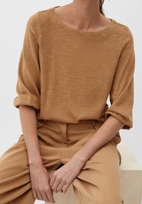 Lässiges, locker sitzendes, braunes Oberteil mit Struktur und weitem Ausschnitt, kombiniert mit passenden beigen Hosen, die ein elegantes, minimalistisches Design zeigen.