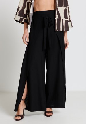 Femme portant un pantalon large noir avec des fentes à l'avant et une ceinture nouée à la taille, associé à des sandales à talons brunes et une veste courte à motifs.