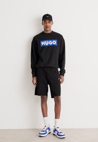 Μαύρη μπλούζα τύπου crewneck με μπλε λογότυπο "HUGO", μαύρες βερμούδες, λευκές κάλτσες και αθλητικά παπούτσια με μπλε λεπτομέρειες. Το μοντέλο στέκεται σε μια απλή φόντο.