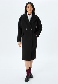 Abrigo de mezcla de lana negro, de doble botonadura, con cuello con muescas, bolsillos frontales y diseño hasta la rodilla. Combinado con calcetines burdeos y mocasines negros.