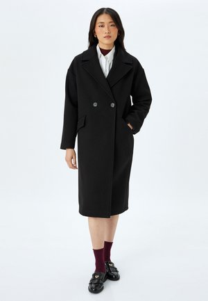 Manteau noir en mélange de laine à double boutonnage avec un col cranté, des poches avant et une longueur jusqu'au genou. Associé à des chaussettes bordeaux et des mocassins noirs.
