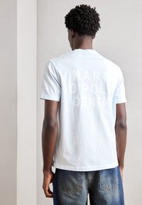 Světle modré bavlněné tričko s krátkými rukávy a kulatým výstřihem. Na zadní straně je velký bílý text: "MARC O'POLO DENIM." Volný střih.
