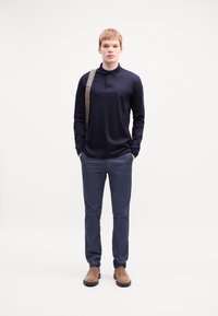 Mornarsko dolgi polo majica, slim-fit temni hlače, svetlo rjavi ante čevlji do gležnja in rjava torba na trak, v stoječem položaju ob enobarvni ozadju.