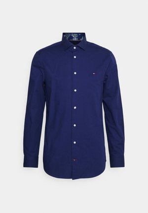 Laivastonsininen pitkiä hihoja oleva nappikauluspaita, jossa on klassinen kaulus ja pieni kirjailtu logo rinnassa, kukkakuvioinen kauluksen sisäpuoli.