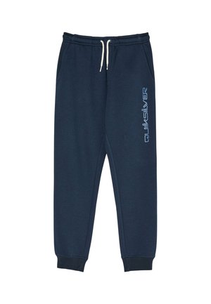 Kind mit lockigem Haar in einem marineblauen Hoodie und passenden Jogginghosen, die Hände in den Taschen, steht vor einem schlichten Hintergrund.