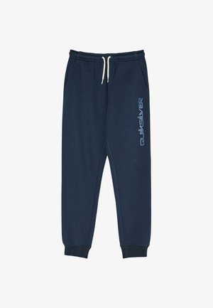Kind mit lockigem Haar in einem marineblauen Hoodie und passenden Jogginghosen, die Hände in den Taschen, steht vor einem schlichten Hintergrund.