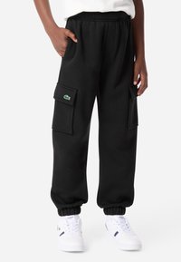 Čierne cargo nohavice s elastickým pásom a manžetami. Obsahujú dve bočné vrecká a zelenú nášivku s logom Lacoste na ľavej nohe.