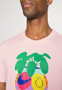 Camiseta de algodón rosa con un colorido estampado gráfico de una planta en una maceta, con hojas verdes y una cara sonriente, sobre un fondo multicolor.