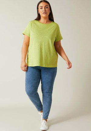 Frau trägt limegrünes kurzärmeliges Oberteil mit kleinen Verzierungen, blaue enge Jeans und weiße Turnschuhe, geht vorwärts vor einfarbigem Hintergrund.