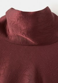 Bordeauxrotes Oberteil mit hohem Kragen, sichtbarer Stoffstruktur und dezenten Falten vor schlichtem weißem Hintergrund.