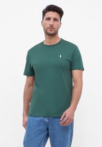 Groene katoenen T-shirt met een ronde halslijn, korte mouwen en een klein wit boomlogo op de borst. Draagt met lichtblauwe spijkerbroek.