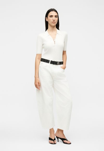 Γυναίκα που στέκεται με το χέρι στην τσέπη φορώντας άσπρο τοπ με κοντά μανίκια, φαρδιά λευκά παντελόνια, μαύρη ζώνη και μαύρα σανδάλια με τακούνι σε λευκό φόντο.