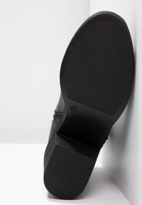 Chaussures noires avec une semelle en caoutchouc texturé, présentant un embout arrondi et des détails de couture sur les côtés. Taille indiquée comme 37 au-dessous.