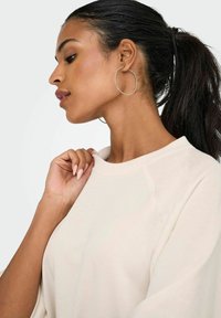 Eine Frau mit dunklem, zurückgebundenem Haar trägt große Creolen und ein hellbeiges Langarmshirt, das sie mit einer Hand am Kragen hält.