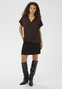 Braune Kurzarmbluse mit V-Ausschnitt, kombiniert mit einem schwarzen, taillierten Rock und glänzenden schwarzen kniehohen Stiefeln mit Reptilstruktur.