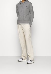 Persona che indossa un hoodie grigio con un piccolo logo a forma di cuore, pantaloni larghi crema e sneakers bianche, in piedi contro un muro bianco.