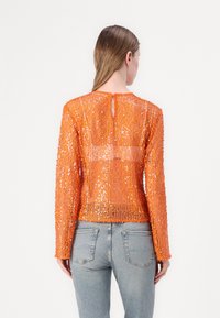 Oranje pailletten top met lange mouwen, gestructureerd oppervlak, sleutelgat sluiting aan de achterkant, gecombineerd met lichtblauwe jeans.