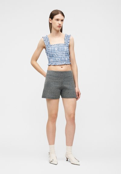 Femme aux cheveux bruns lisses portant un haut court sans manches à carreaux bleus, un short gris, des chaussettes crème et des chaussures blanches à talons, debout devant un fond blanc.