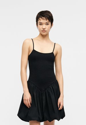 Vrouw met kort donker haar draagt een mouwloze zwarte jurk met een nauwsluitend lijfje en geplooide ballerina-rok, staand tegen een effen lichte achtergrond.