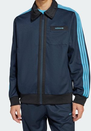 Mann trägt dunkelblauen Adidas Trainingsanzug mit hellblauen Streifen und Logo, steht mit entspannten Händen seitlich am Körper.