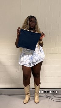 Mujer con cabello largo y rubio que lleva una falda blanca y azul y botas beige, sosteniendo un cuadrado de tela azul marino contra una pared lisa.