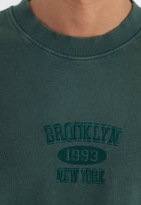 Donkergroen sweatshirt met geribbelde ronde hals. Bevat geborduurde tekst: "BROOKLYN," "1993," en "NEW YORK" in verschillende tinten groen.