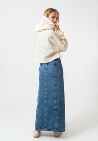 Kremasti kratki dukserica s prednjim džepom i vezenim detaljima, u kombinaciji s plavom jeans suknjom maxi koja ima vertikalne šavove i ravni kroj.