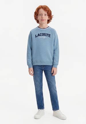 Garçon aux cheveux roux portant un sweat Lacoste bleu clair, un jean bleu et des baskets blanches, debout devant un fond blanc.