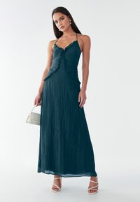 Abito maxi verde acqua con una trama texturizzata e pieghettata, caratterizzato da bordi arricciati e sottili spalline a spaghetti. La modella tiene in mano una piccola borsa bianca.
