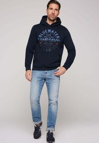 Sudadera navy con un bolsillo canguro frontal, con texto gráfico en azul y rojo. Combinada con jeans de un azul claro y zapatillas oscuras.