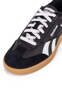 Adidași din piele neagră cu șireti albi, având un vârf din suede și o talpă din cauciuc căprui. Include accente laterale albe și negre.