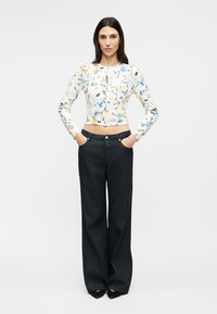 Vrouw met stijl donker haar draagt een bloemenprint cropped top met lange mouwen en hooggesloten, wijde zwarte broek met handen in de zakken.