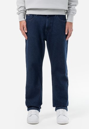 Dark blue denim jeans with a relaxed fit, featuring five pockets, and subtle stitching details, paired with white sneakers.