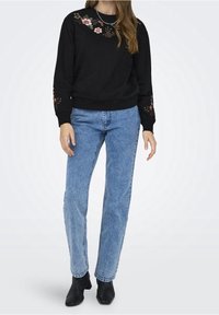 Sudadera negra con bordado floral en el escote y las mangas, combinada con jeans rectos de color azul claro y botines negros.